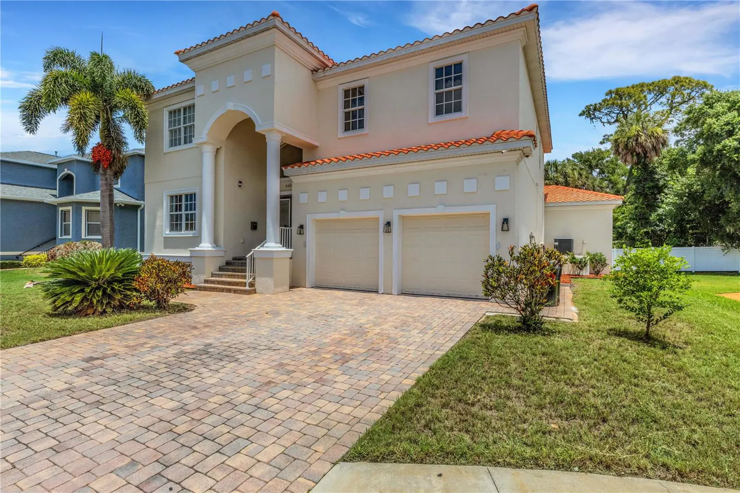 Two-story beige stucco home with a terracotta tile roof, two-car garage, and a paved driveway under a bright blue sky.