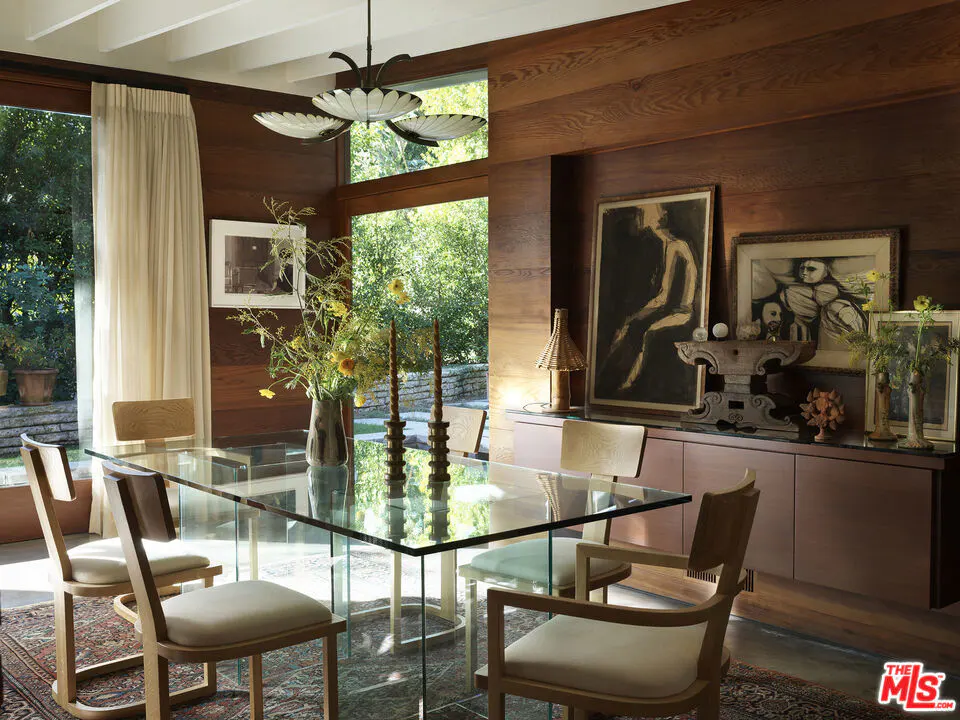 Dining room with wood paneled walls, a glass table, and light wood chairs. Art hangs above a brown cabinet. A window looks out to greenery.