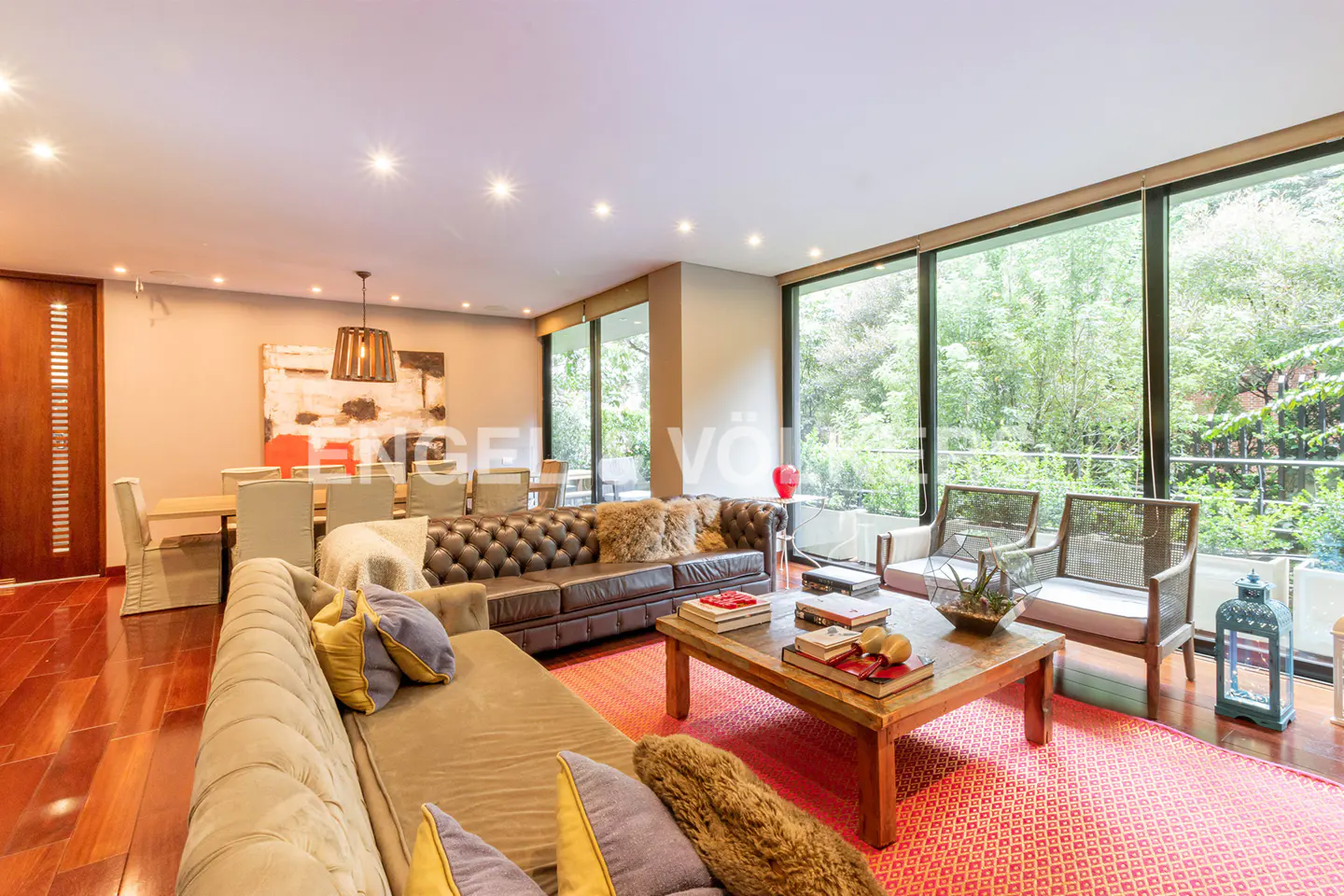 Bright living room with brown leather sofa, wood coffee table, and red patterned rug. Large windows overlook lush greenery. Dining area in background.