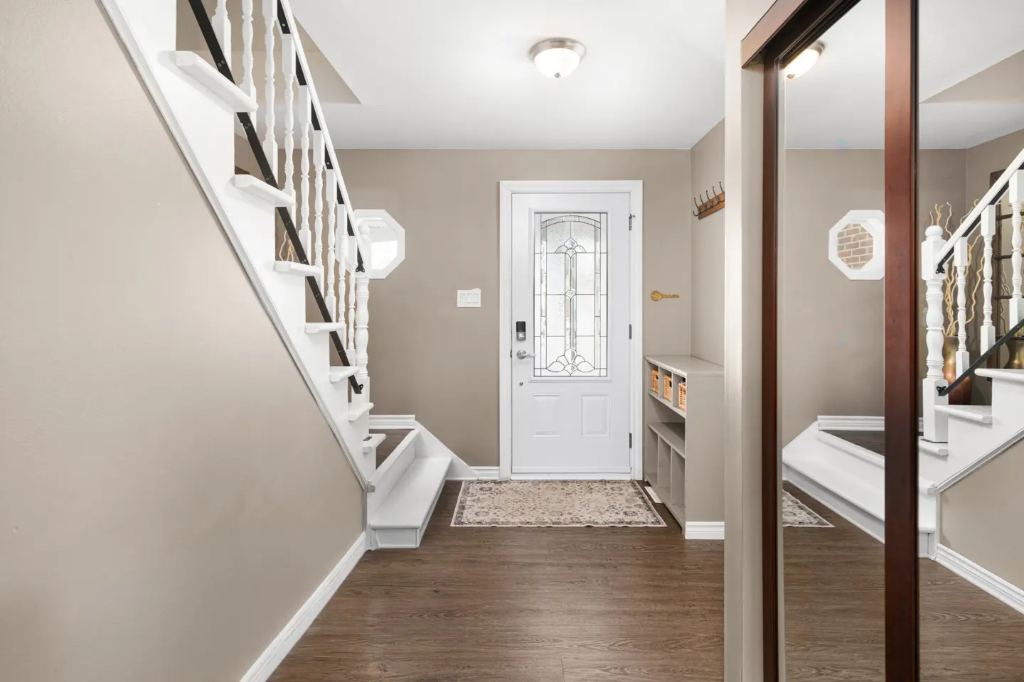 Foyer with white door, stairs, and wood floors. A mirror reflects the staircase. Walls are beige.