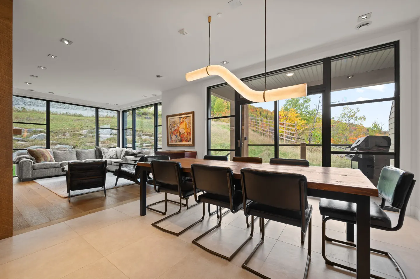 Bright, modern dining area with a long wooden table, black chairs, and a unique, wavy light fixture. Large windows offer scenic outdoor views.