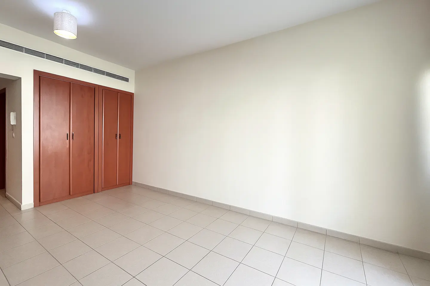Empty room with beige tile floor, cream walls, and a built-in brown wood closet. A white light fixture is on the ceiling.