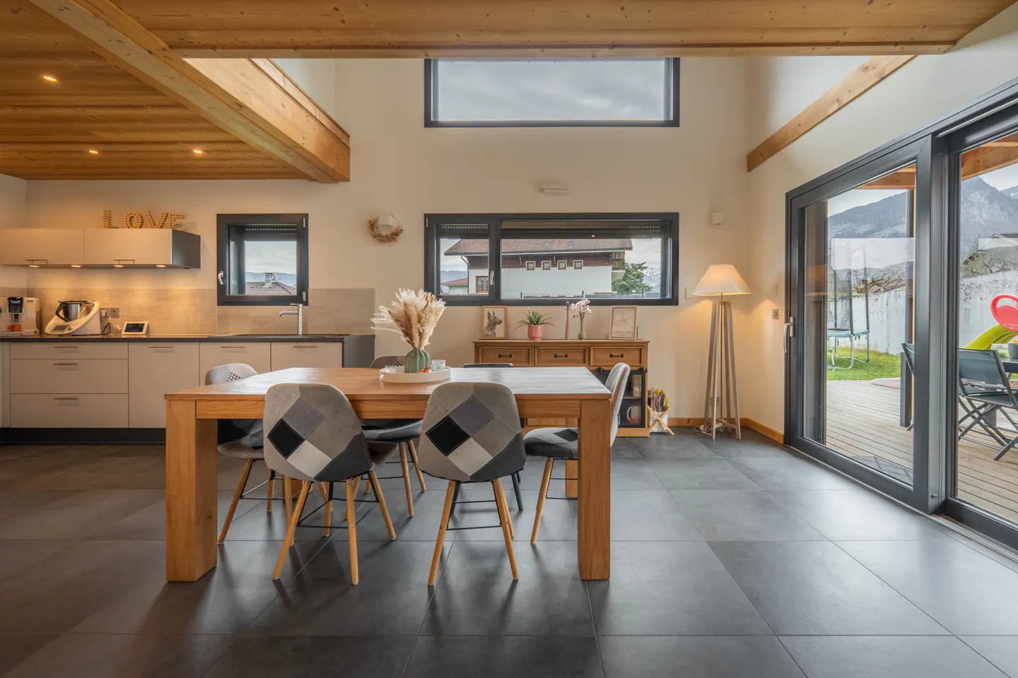 A bright, modern kitchen and dining area with a wooden table, patterned chairs, and large windows overlooking a green yard.