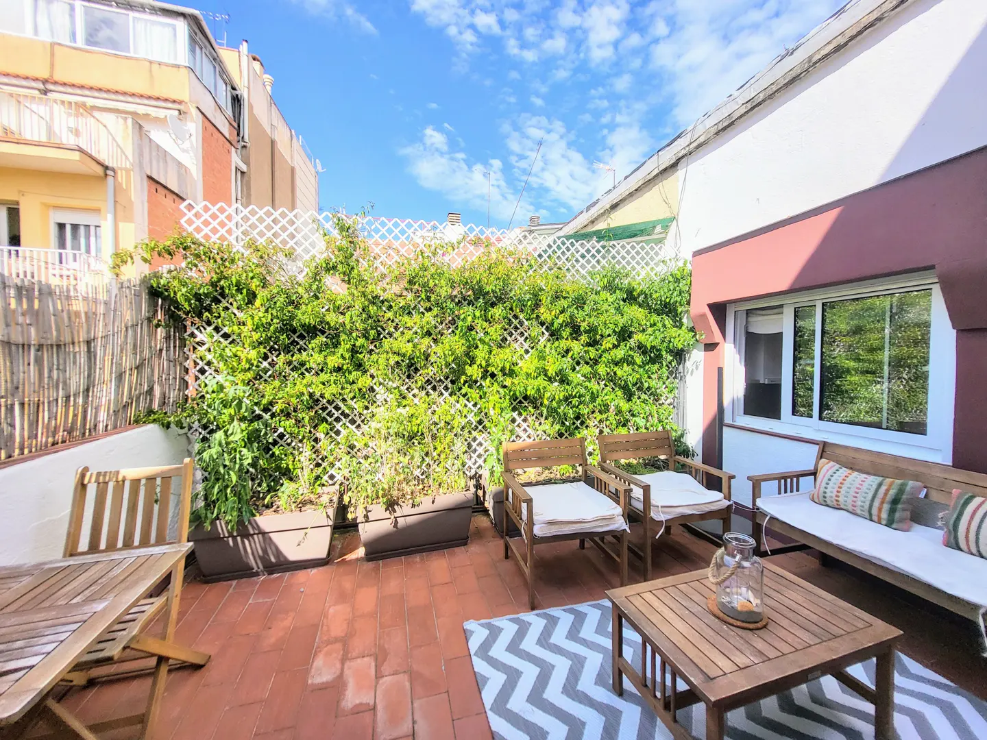 Outdoor patio with brick flooring, wooden furniture, and a white lattice fence covered in green vines. A gray and white chevron rug sits under a wooden coffee table.