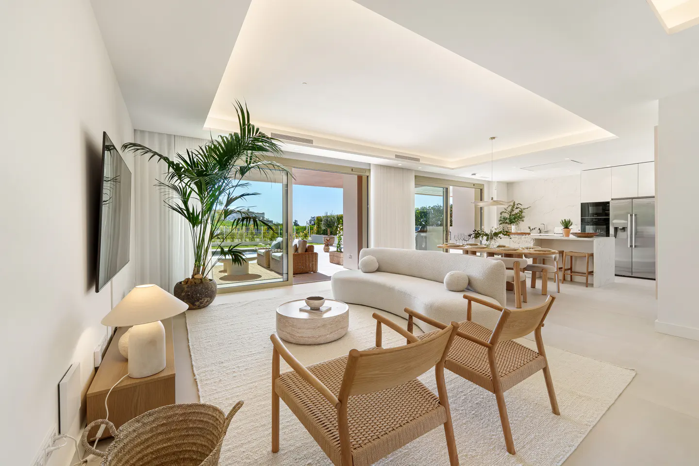 Bright, open-plan living space with white walls, light wood furniture, and a large potted plant. Sliding glass doors lead to an outdoor patio.