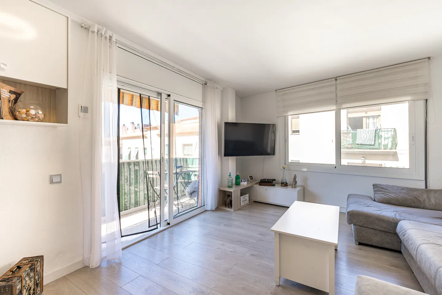 Bright living room with a grey sofa, white coffee table, wall-mounted TV, and sliding glass doors leading to a balcony.