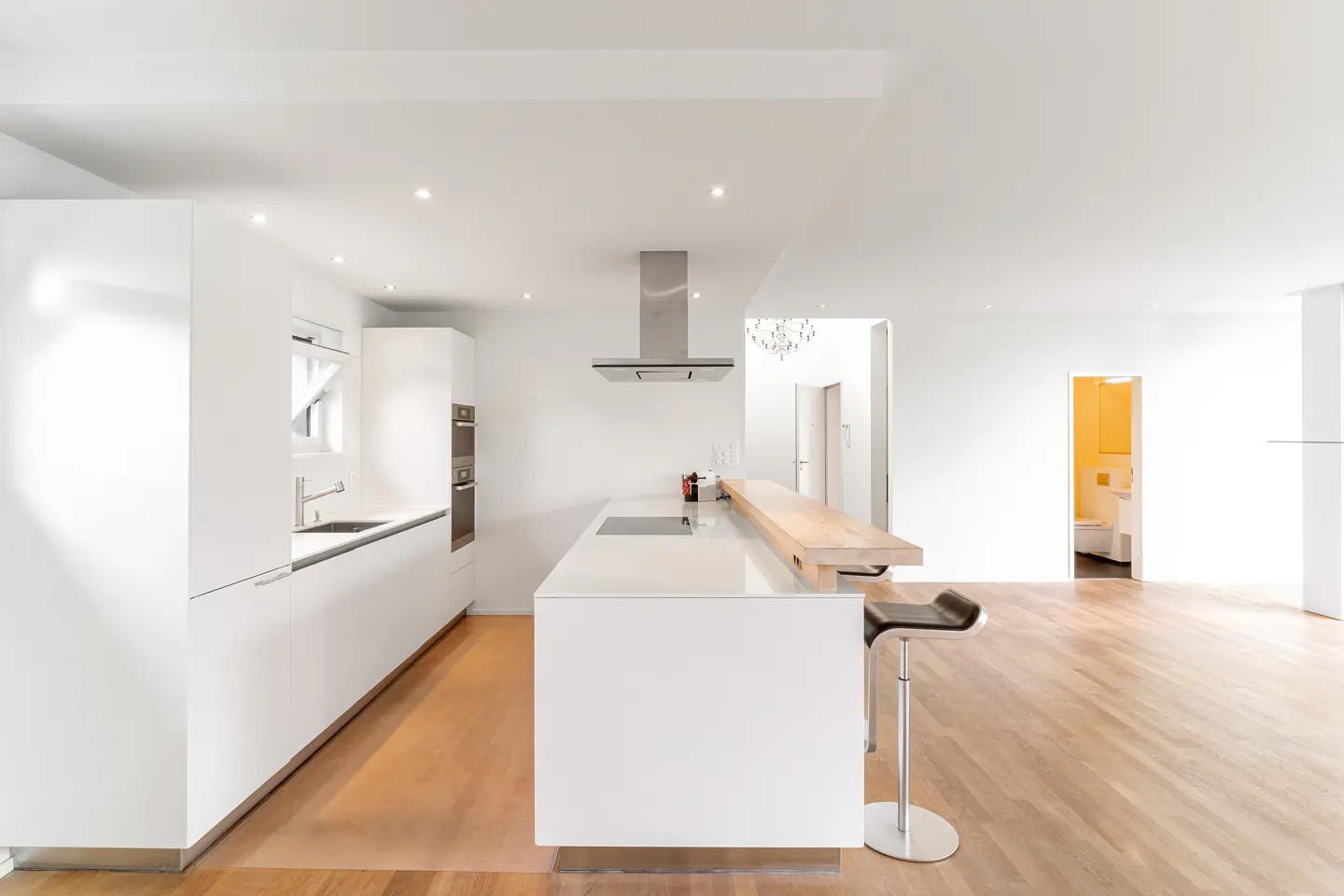 Modern white kitchen with a central island, wooden breakfast bar, stainless steel hood, and light hardwood flooring.
