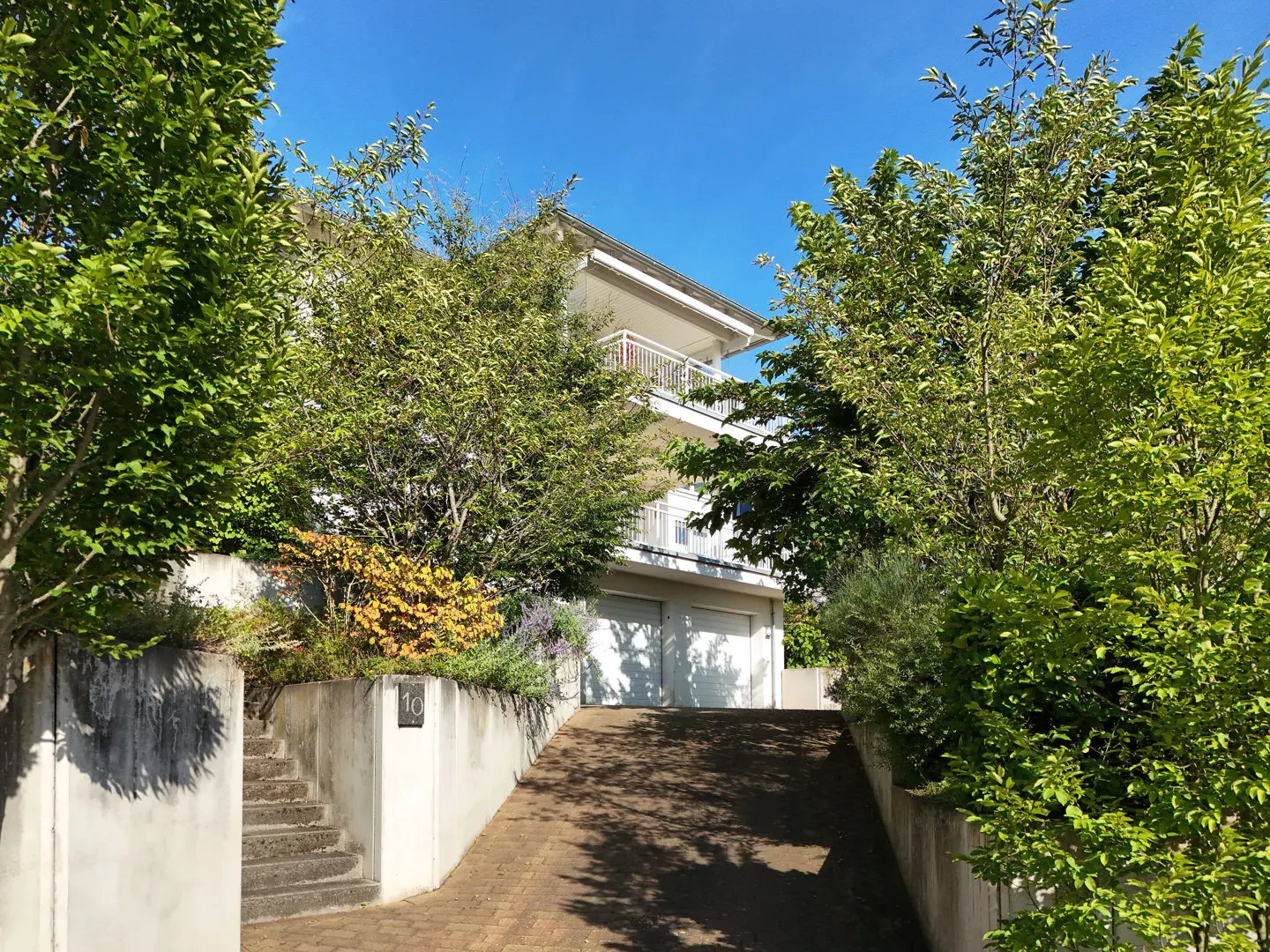 Two-story white house with a balcony, surrounded by green trees, viewed from the driveway. Blue sky.
