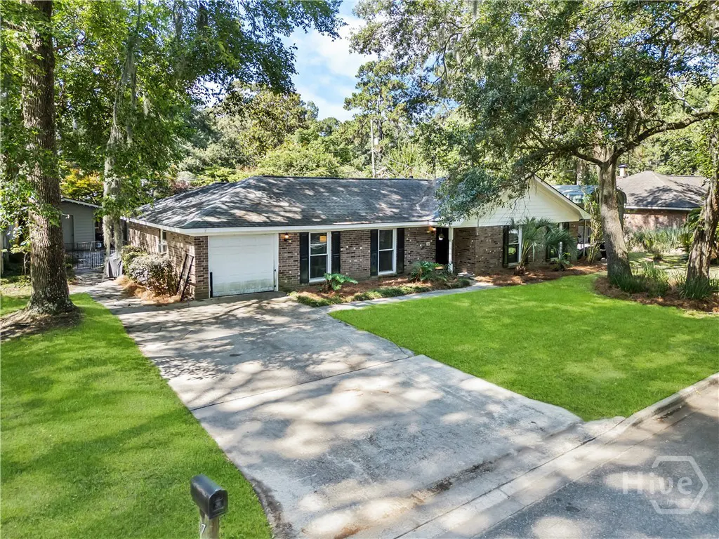 Single-story brick ranch house with a white garage, concrete driveway, and green lawn surrounded by mature trees.