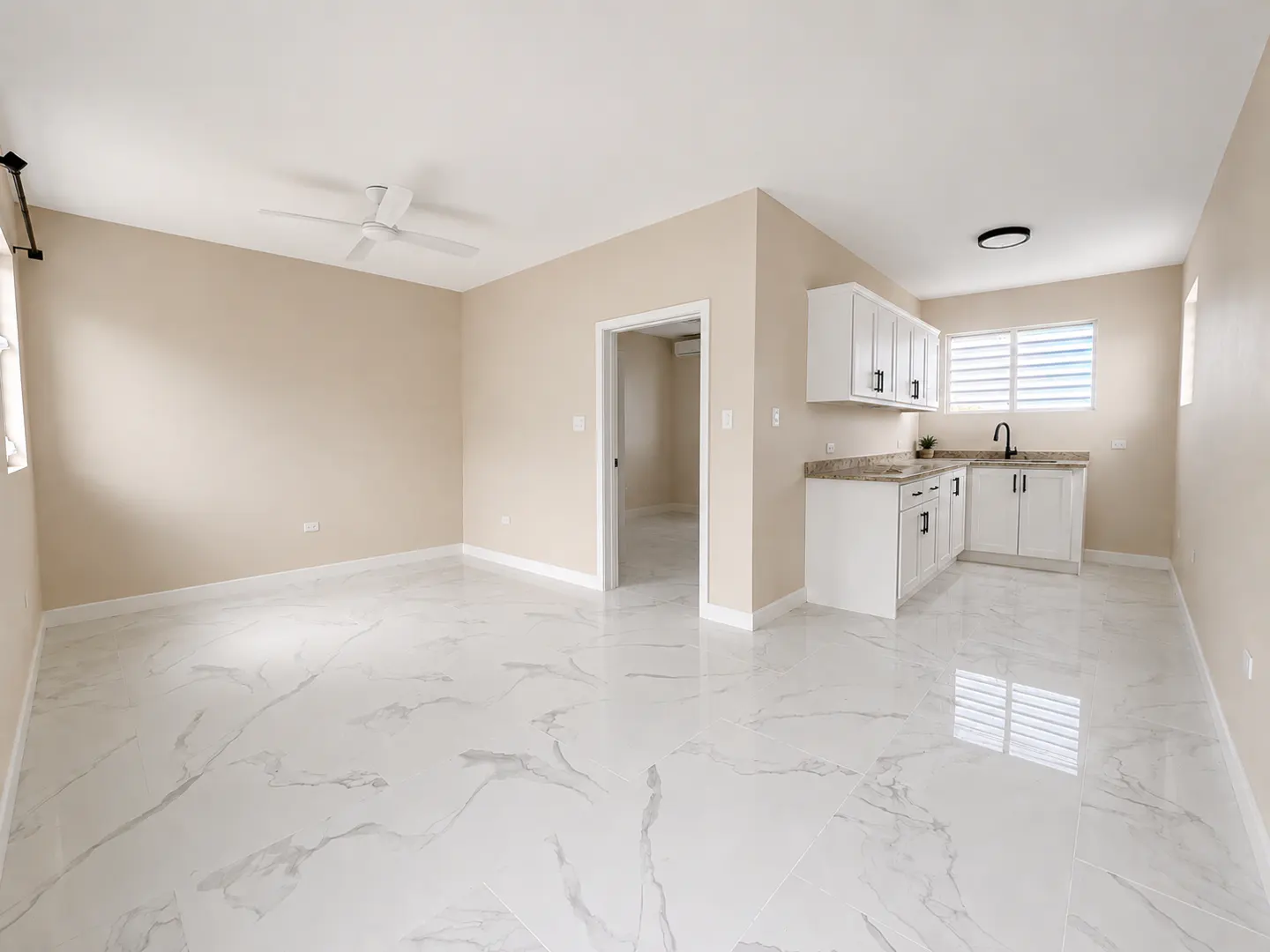 Bright, empty room with white marble floors, beige walls, and a small kitchen area with white cabinets. A doorway leads to another room.