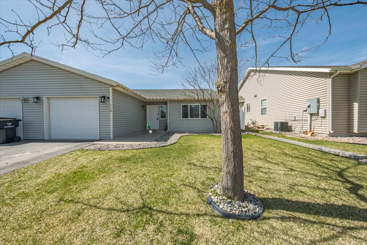 Exterior view of a one-story home with light gray siding, a white garage door, and a green lawn. A tree stands in the yard.