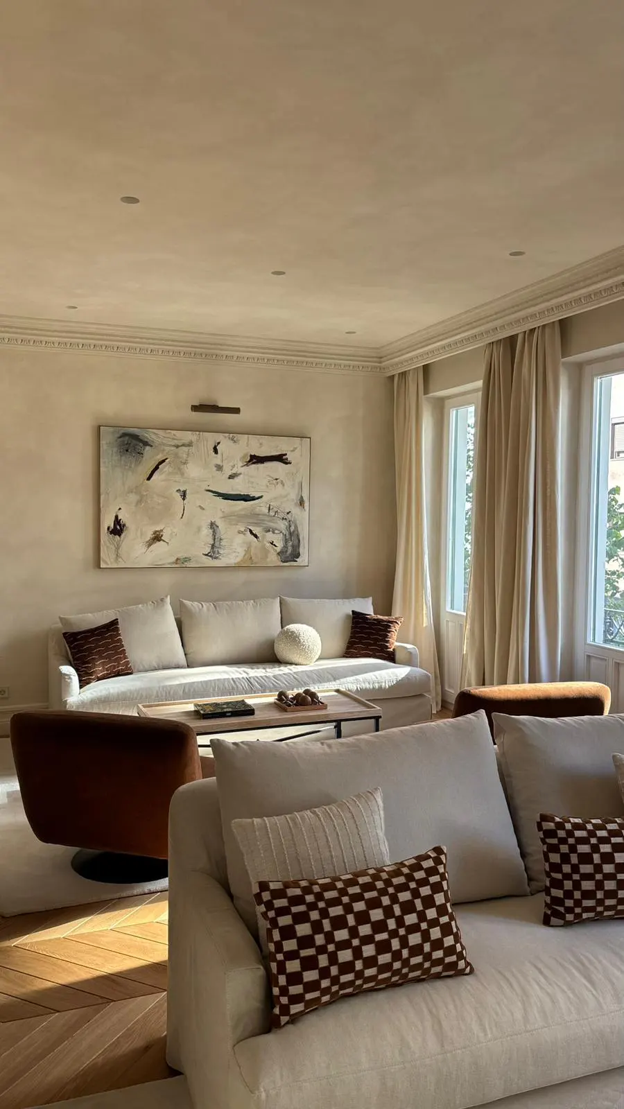 Living room with neutral tones, featuring white sofas with patterned pillows, a brown swivel chair, and an abstract painting.
