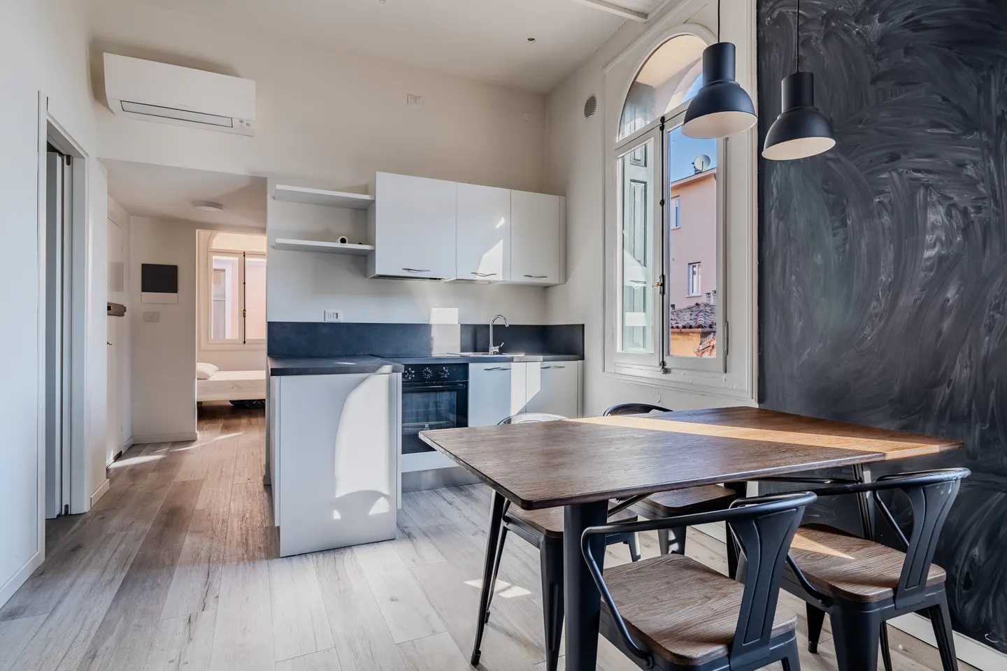 Bright, modern kitchen with white cabinets, dark countertops, and a wooden table with black chairs. A large window overlooks a building.