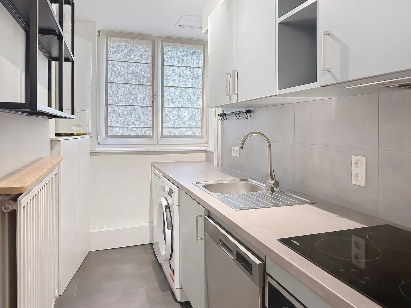 A modern, compact kitchen with white cabinets, stainless steel appliances, and a gray countertop. A window with blinds provides natural light.
