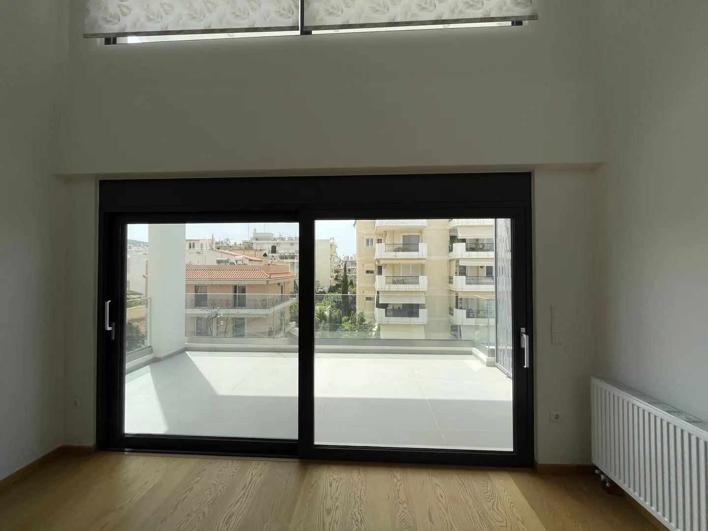 A bright, empty room with light wood floors and a large sliding glass door to a balcony overlooking a cityscape.