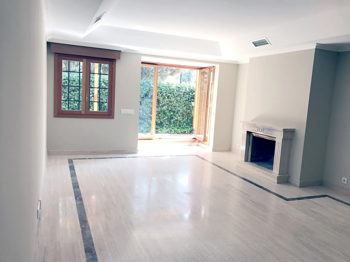 Spacious living room with light marble floors, a stone fireplace, and large windows looking out onto a green hedge.