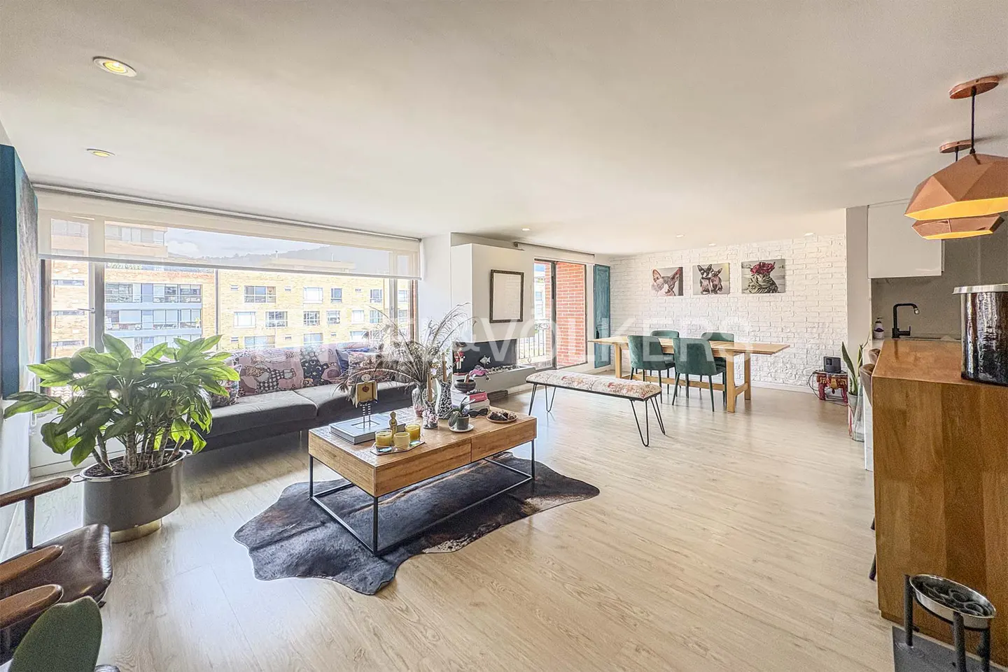 Bright, open-concept living space with a brown sofa, wood coffee table on a cowhide rug, and a dining table with green chairs. Large windows offer city views.