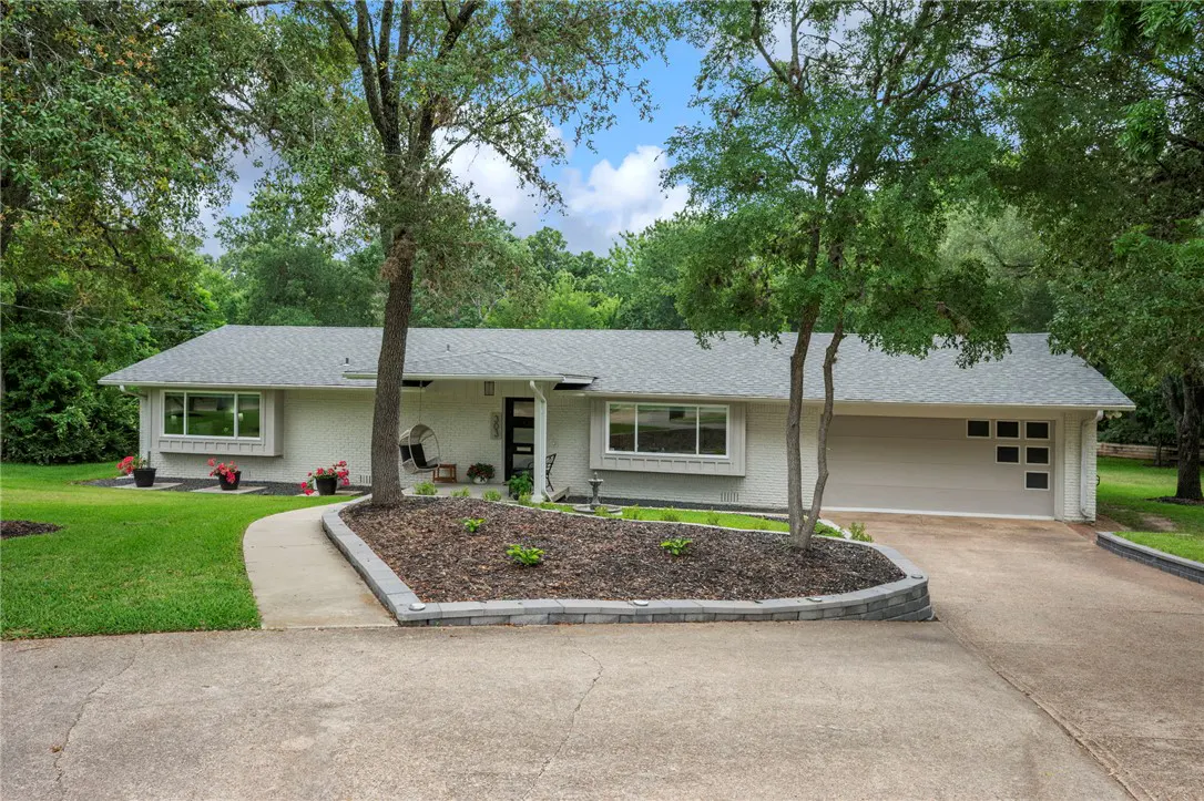 A single-story white house with a gray roof, a two-car garage, and a curved driveway. Trees frame the house.