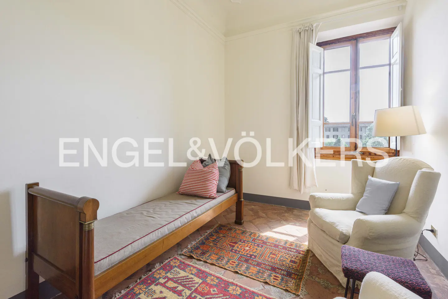 A cozy room with a wooden daybed, a cream armchair, a floor lamp, and patterned rugs on a terracotta tile floor.