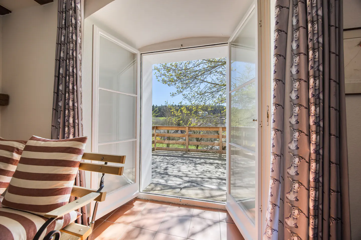 Open white French doors lead to a patio with a wooden fence and trees. Striped pillows sit on a chair. Donkey-patterned curtains frame the doorway.