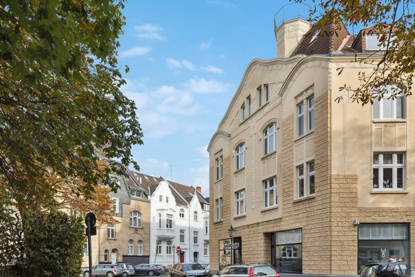 Street view of beige buildings with white trim, cars parked along the curb, and trees with green and yellow leaves.