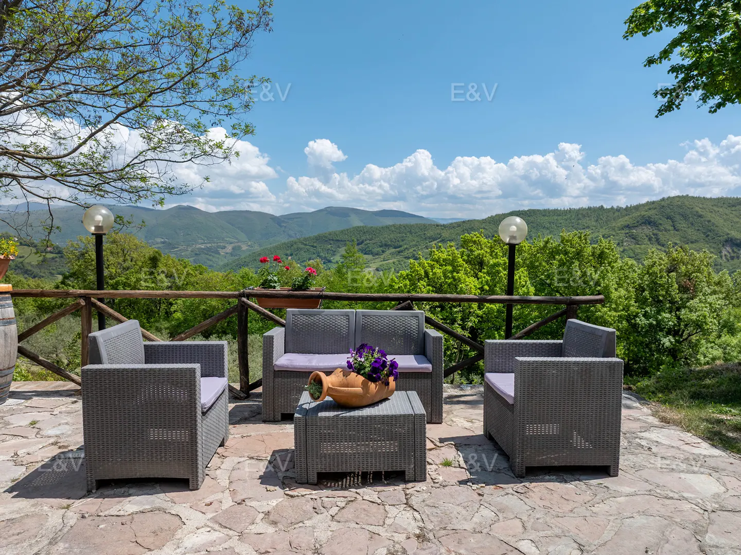 Outdoor patio with gray wicker furniture, purple cushions, and a flower-filled vase, overlooking green hills under a blue sky.