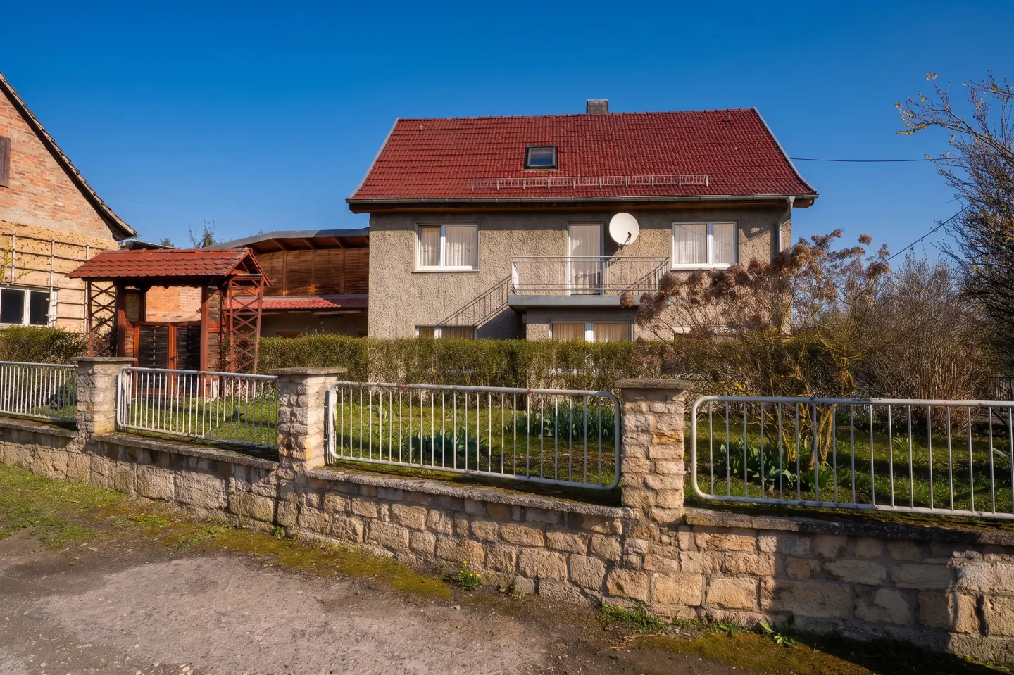 Two-story house with a red tile roof, stone fence, and a small wooden gazebo in the front yard under a clear blue sky.
