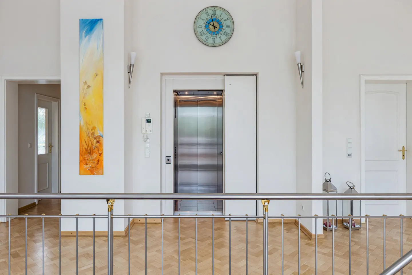 Bright, white hallway with a stainless steel elevator, a blue and yellow painting, and a clock above the elevator.