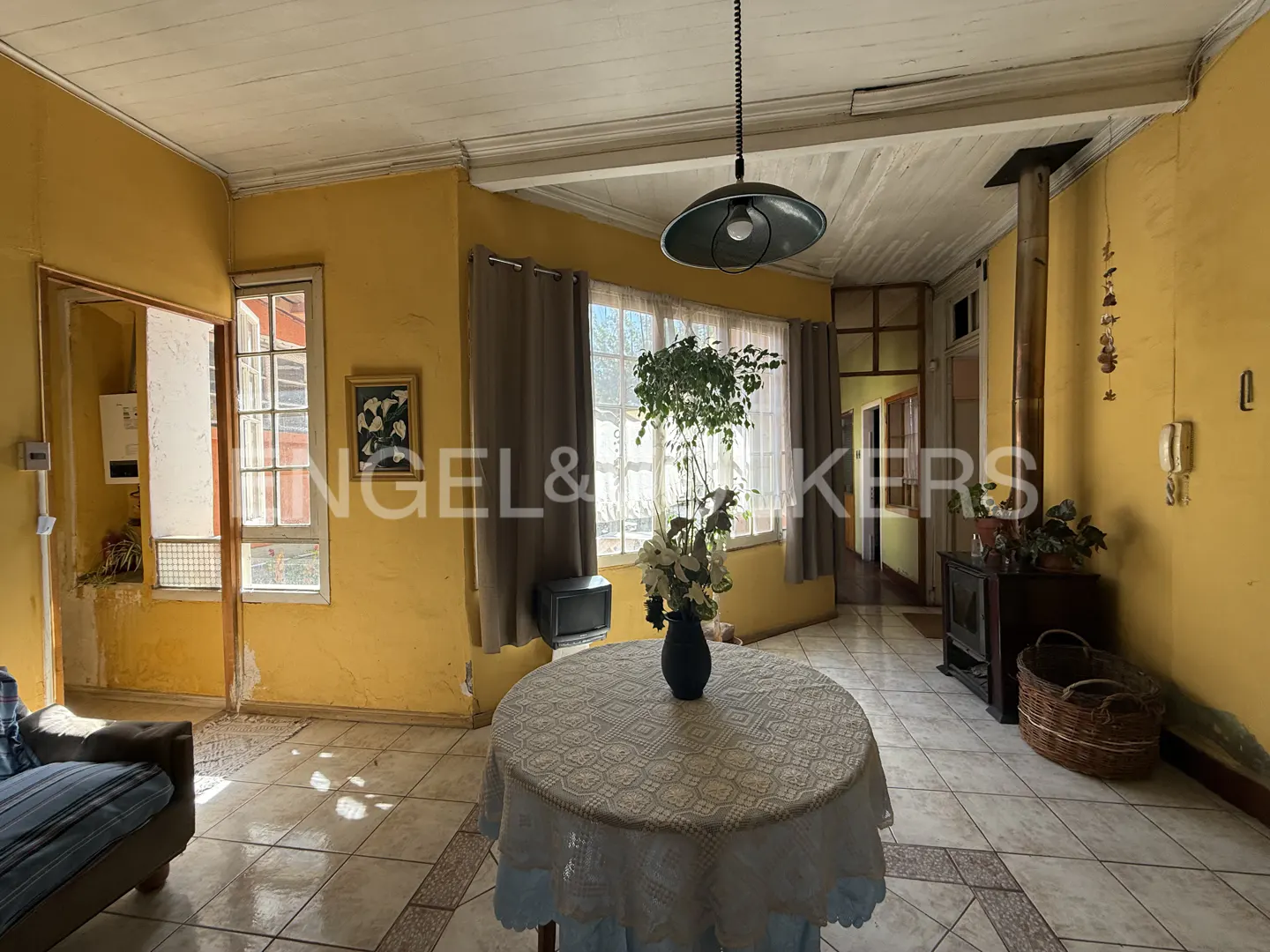 A living room with yellow walls, tiled floors, a round table with a lace tablecloth, and a wood-burning stove.