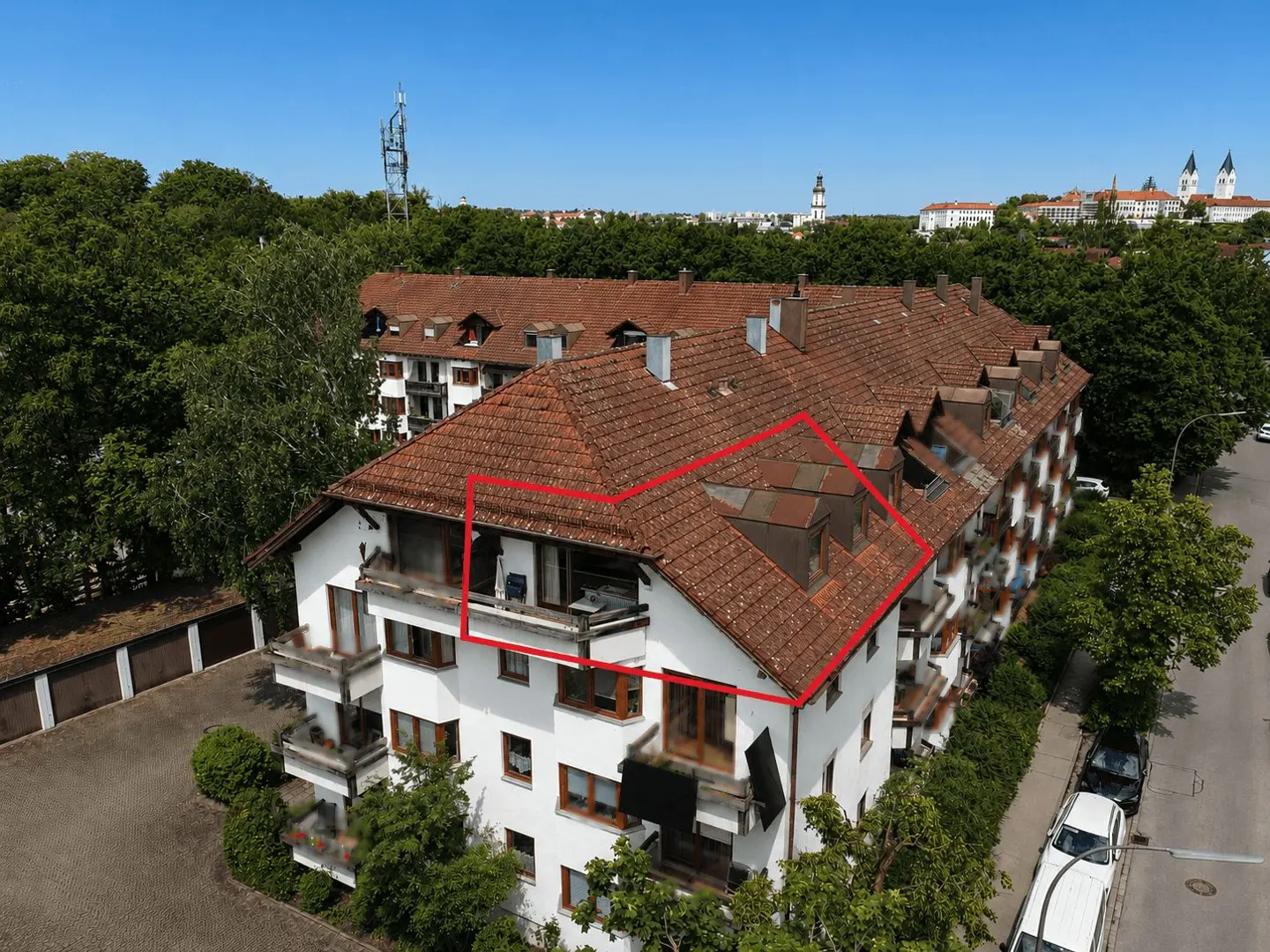 Aerial view of a white apartment building with a red tile roof, highlighting a top-floor unit with a balcony in red outline.