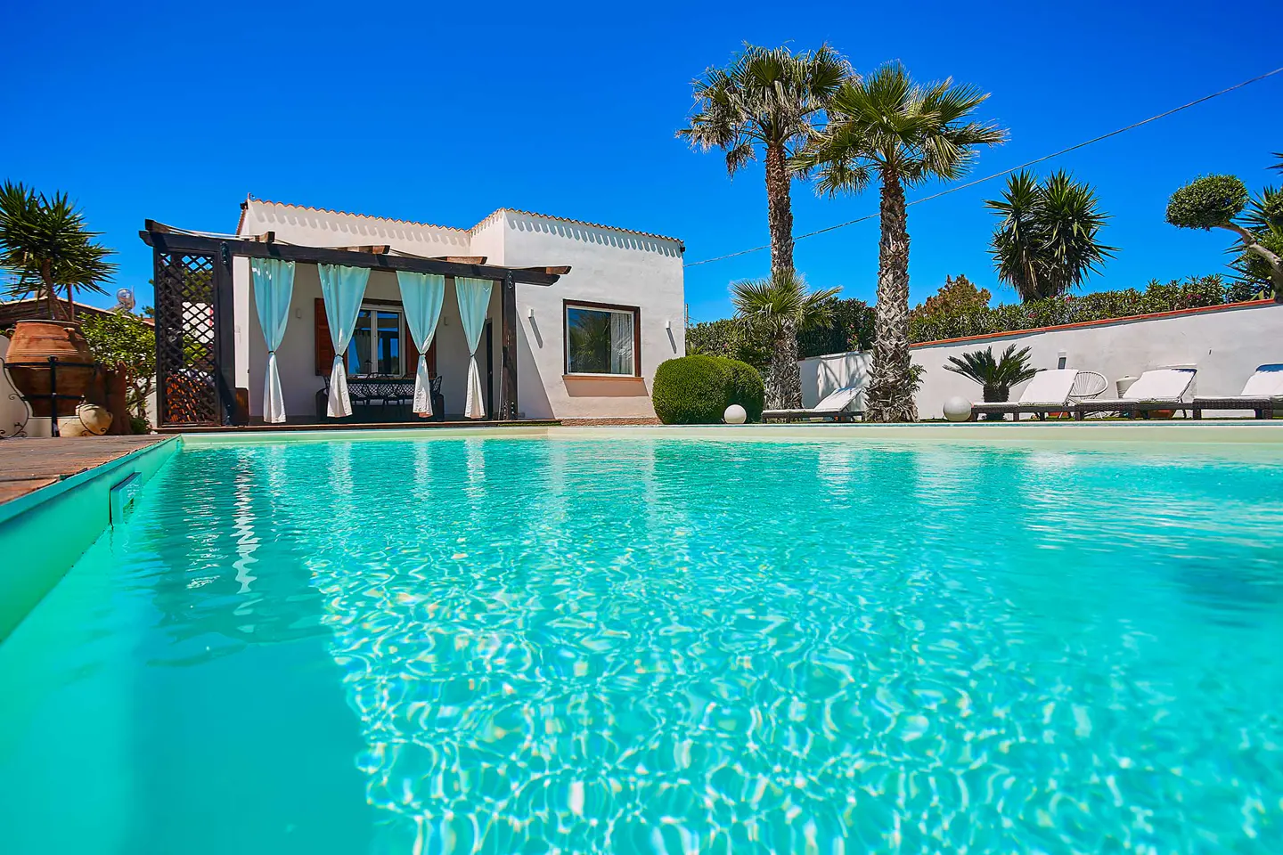 A bright turquoise pool in front of a white house with palm trees under a clear blue sky.