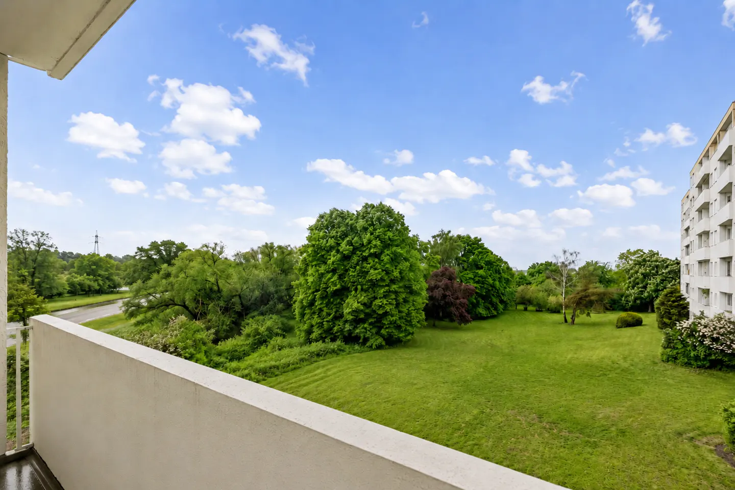 View from a balcony overlooking a green lawn with trees, a road, and a white apartment building under a blue sky with clouds.