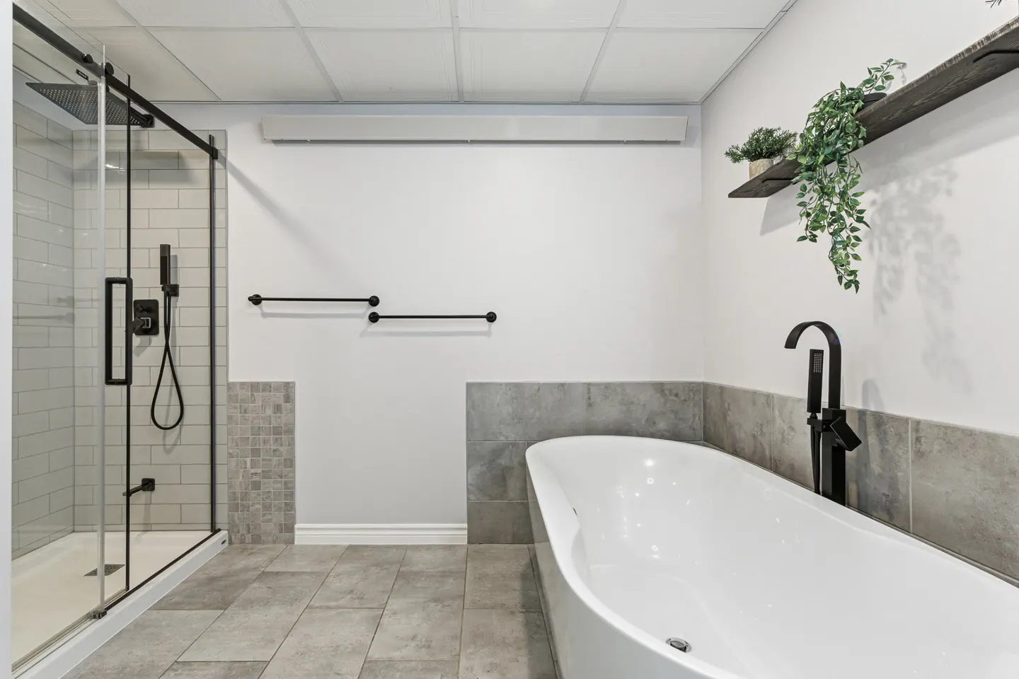 Modern bathroom with a white tub, black faucet, and glass shower. Gray tile floor and white walls. Plants on a shelf add a touch of nature.