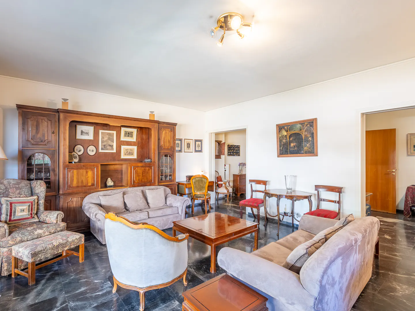 Spacious living room with dark marble floors, two grey sofas, patterned armchairs, and a wooden coffee table.