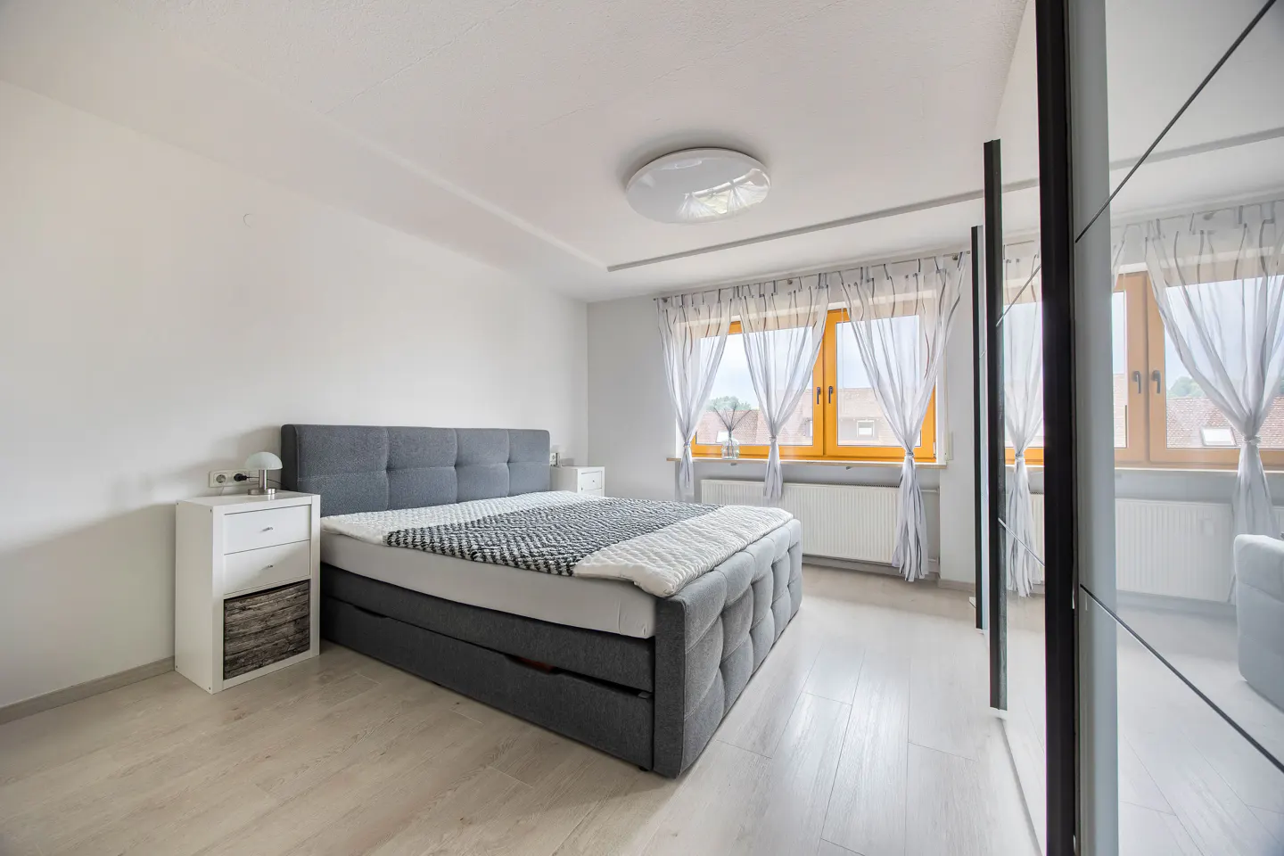 Bright bedroom with a gray upholstered bed, white walls, and light wood floors. A white nightstand sits beside the bed. Windows with sheer curtains let in natural light.
