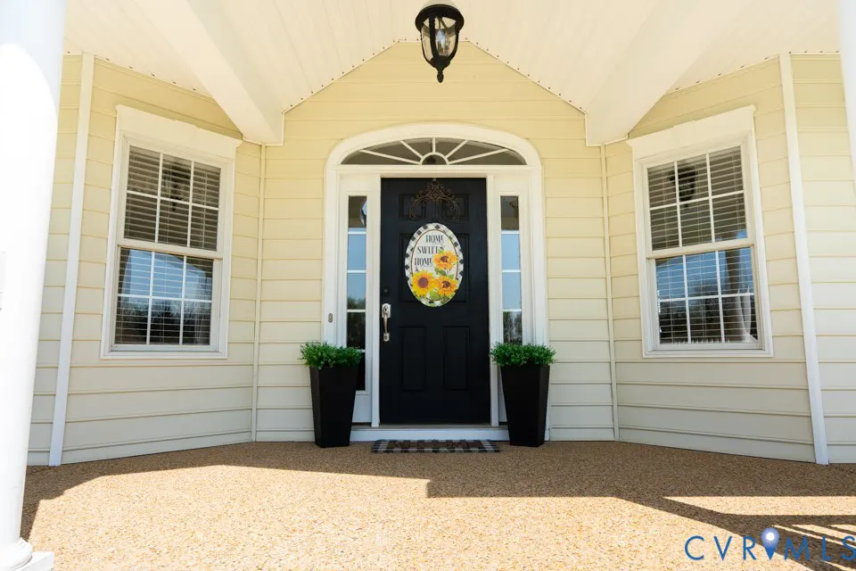 Front exterior of a light yellow house with a black door and "Home Sweet Home" sunflower wreath. Two black planters flank the door.