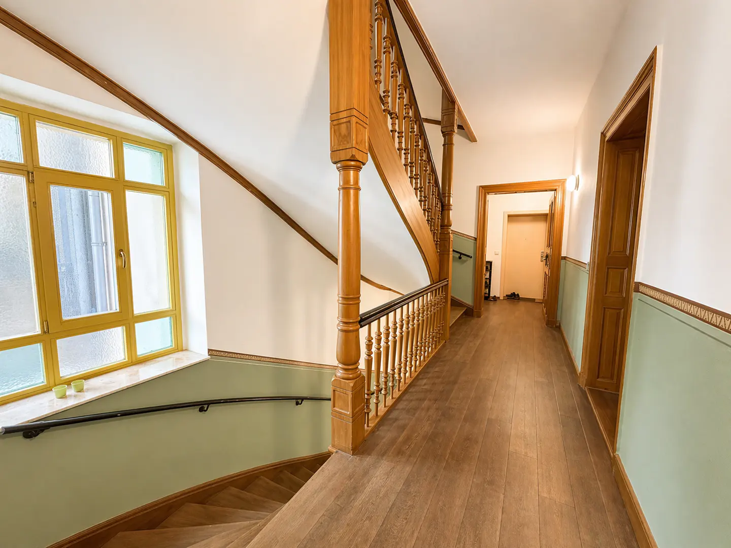 Hallway with wooden floors, stairs, and a yellow-framed window. Walls are white and light green. A wooden banister lines the staircase.