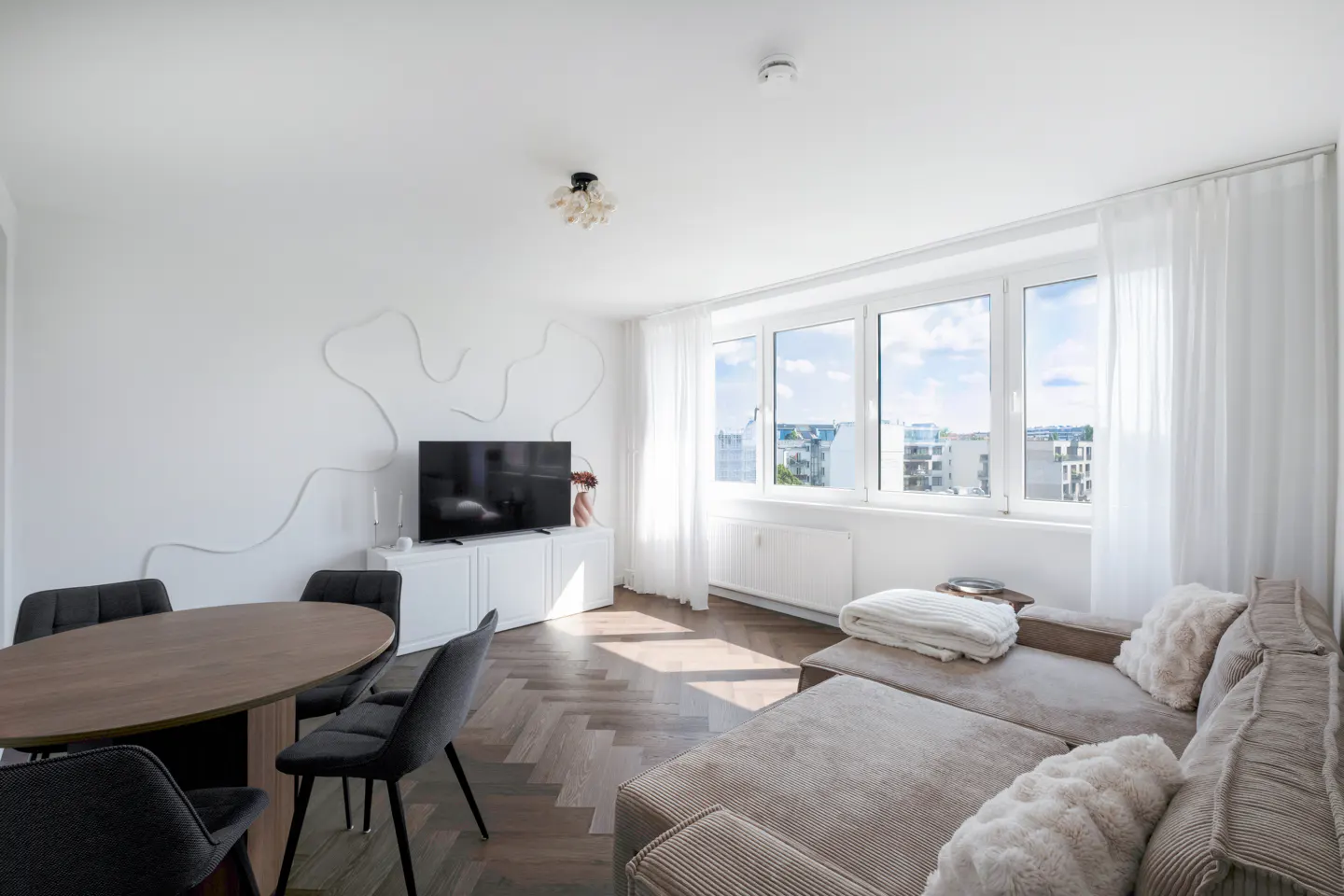 Bright living room with a brown oval table, gray chairs, a beige sectional sofa, and a large window with white curtains.