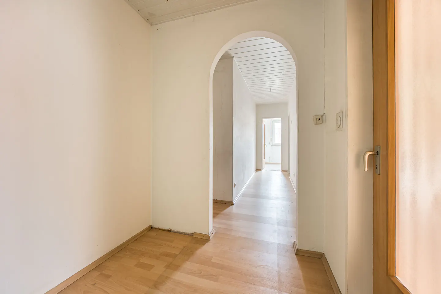 A bright hallway with light wood flooring, white walls, and an arched doorway leading to a sunlit room.
