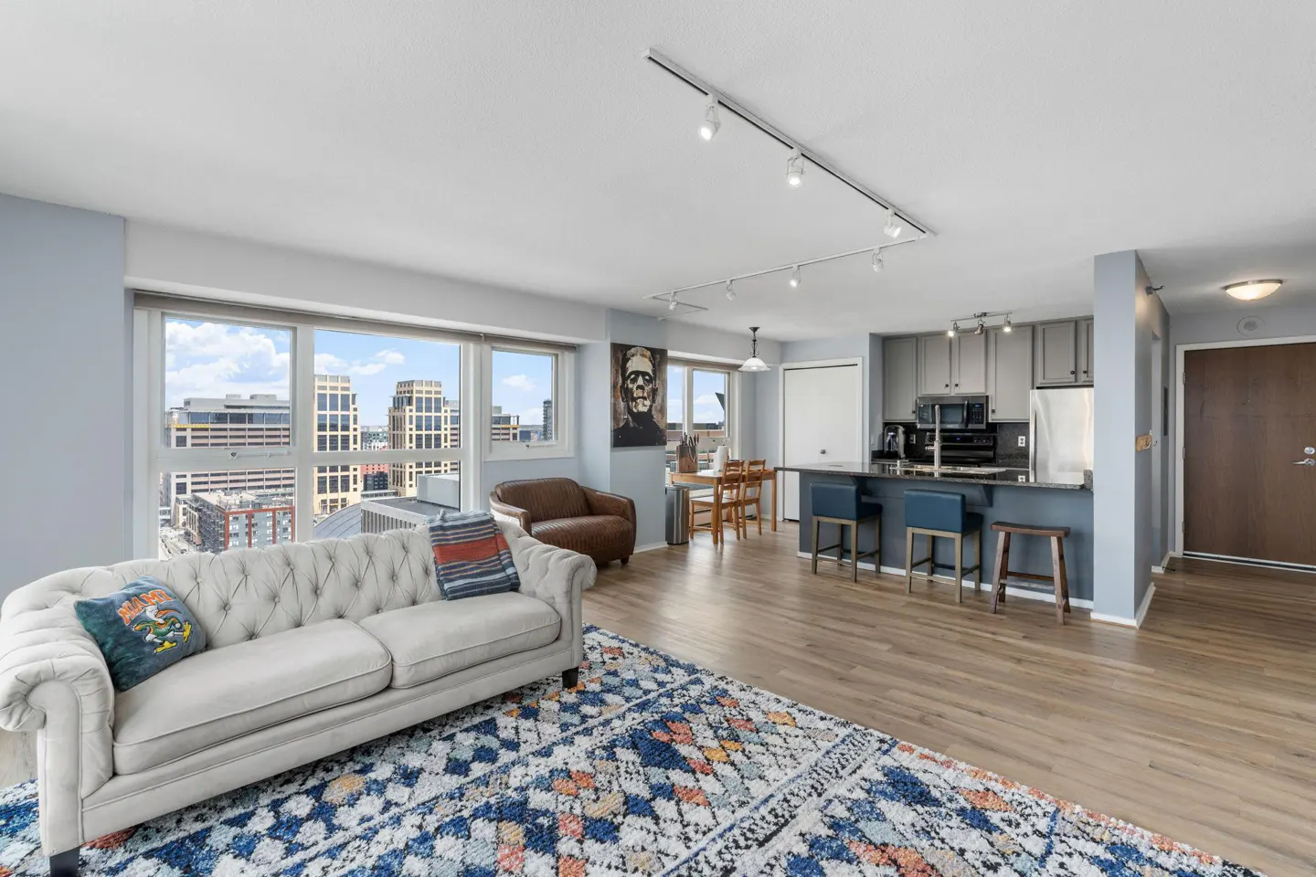 Open-concept apartment with a white sofa, blue rug, and city views from the large windows. The kitchen has gray cabinets and a blue island.