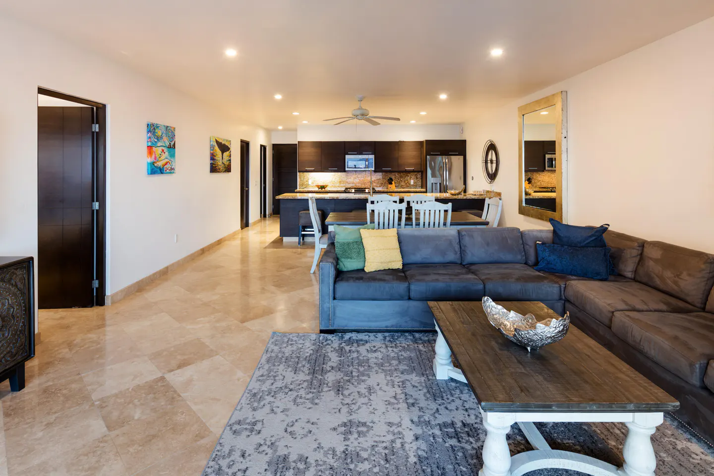 Open-concept living space with a gray sectional sofa, wood coffee table, and kitchen with dark cabinets.