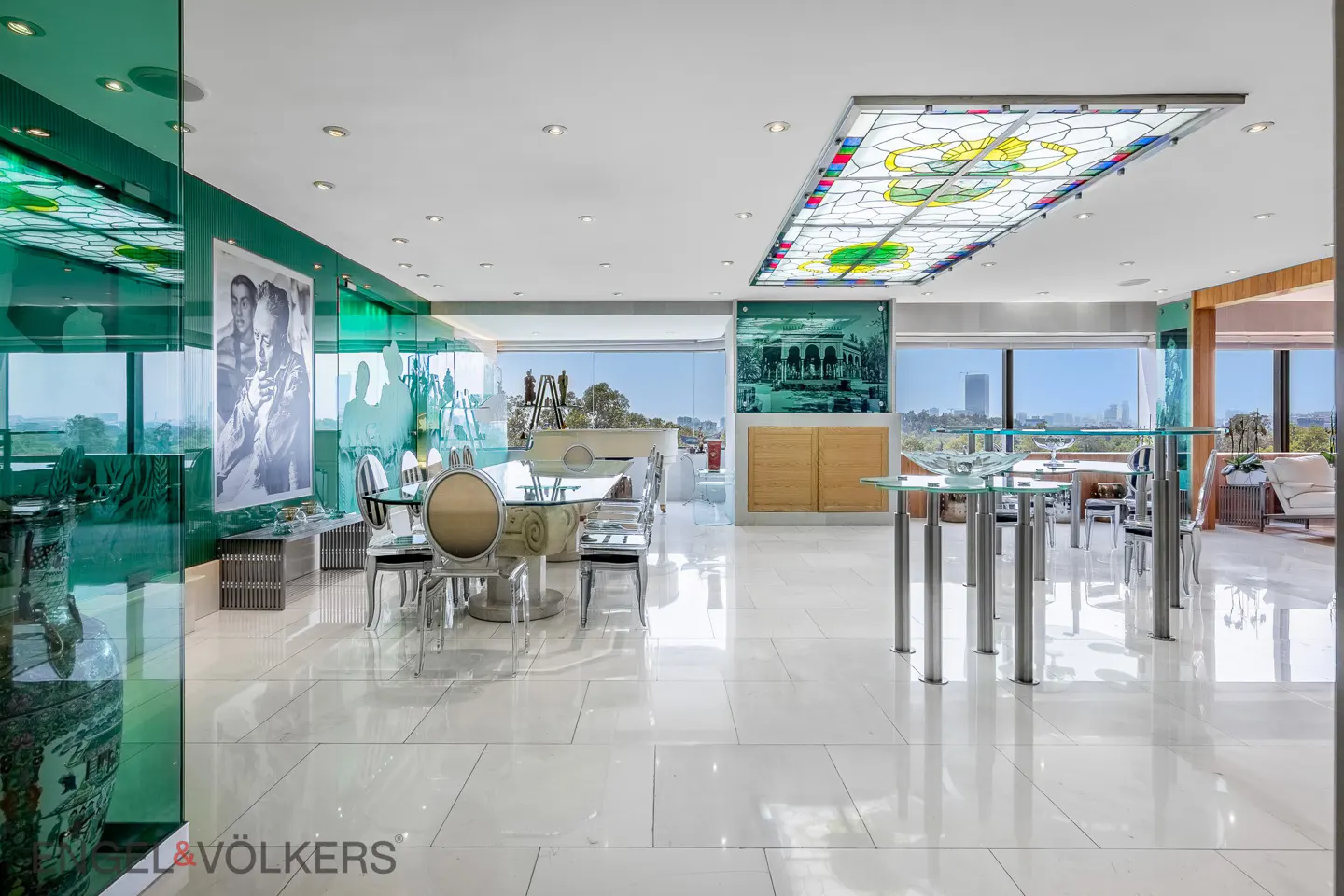 Bright, modern dining room with white tile floors, glass tables, and clear chairs. A stained glass ceiling panel adds color. Large windows offer a city view.