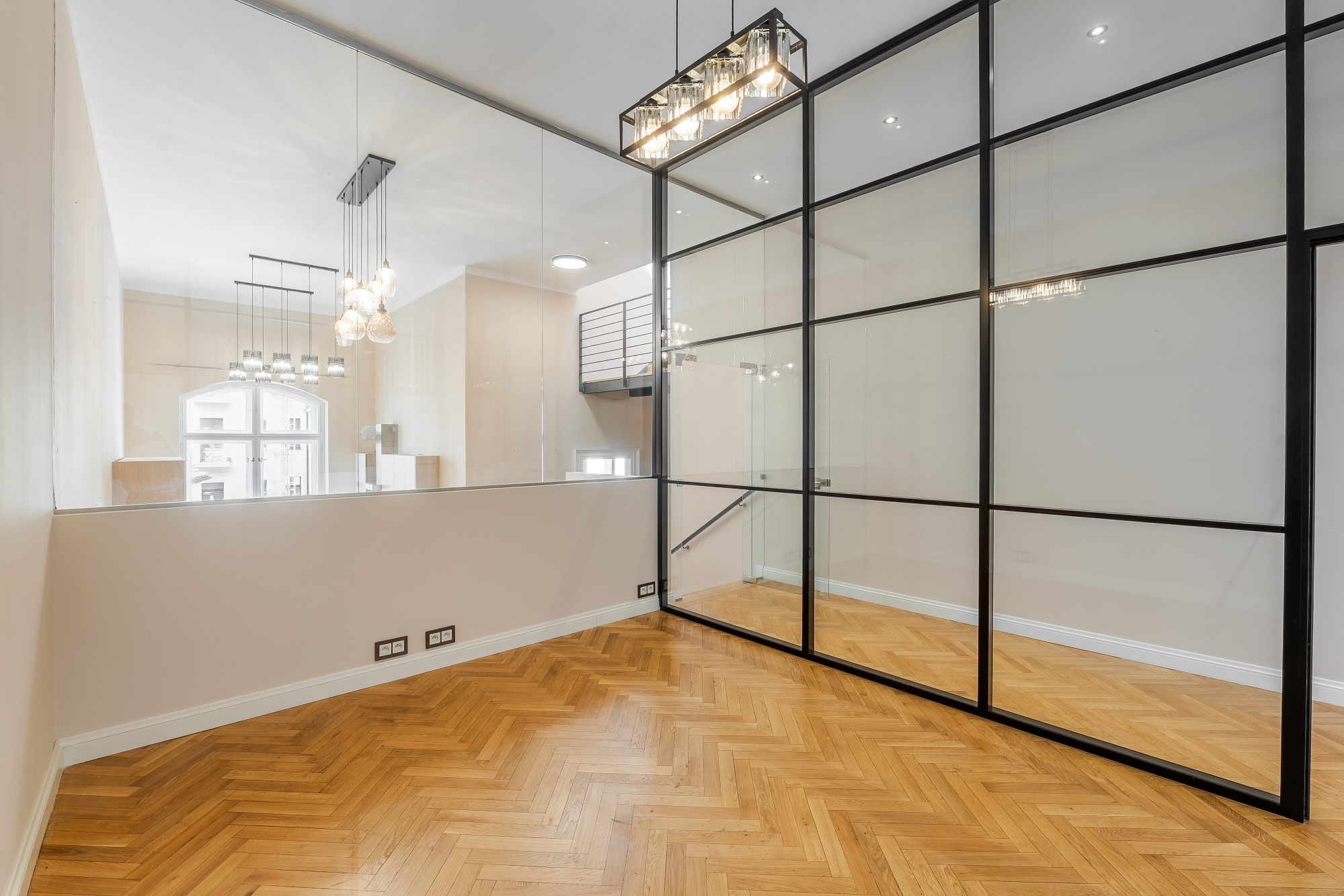 Bright room with herringbone wood floors, a black-framed glass wall, and a partial wall overlooking another room with pendant lights.