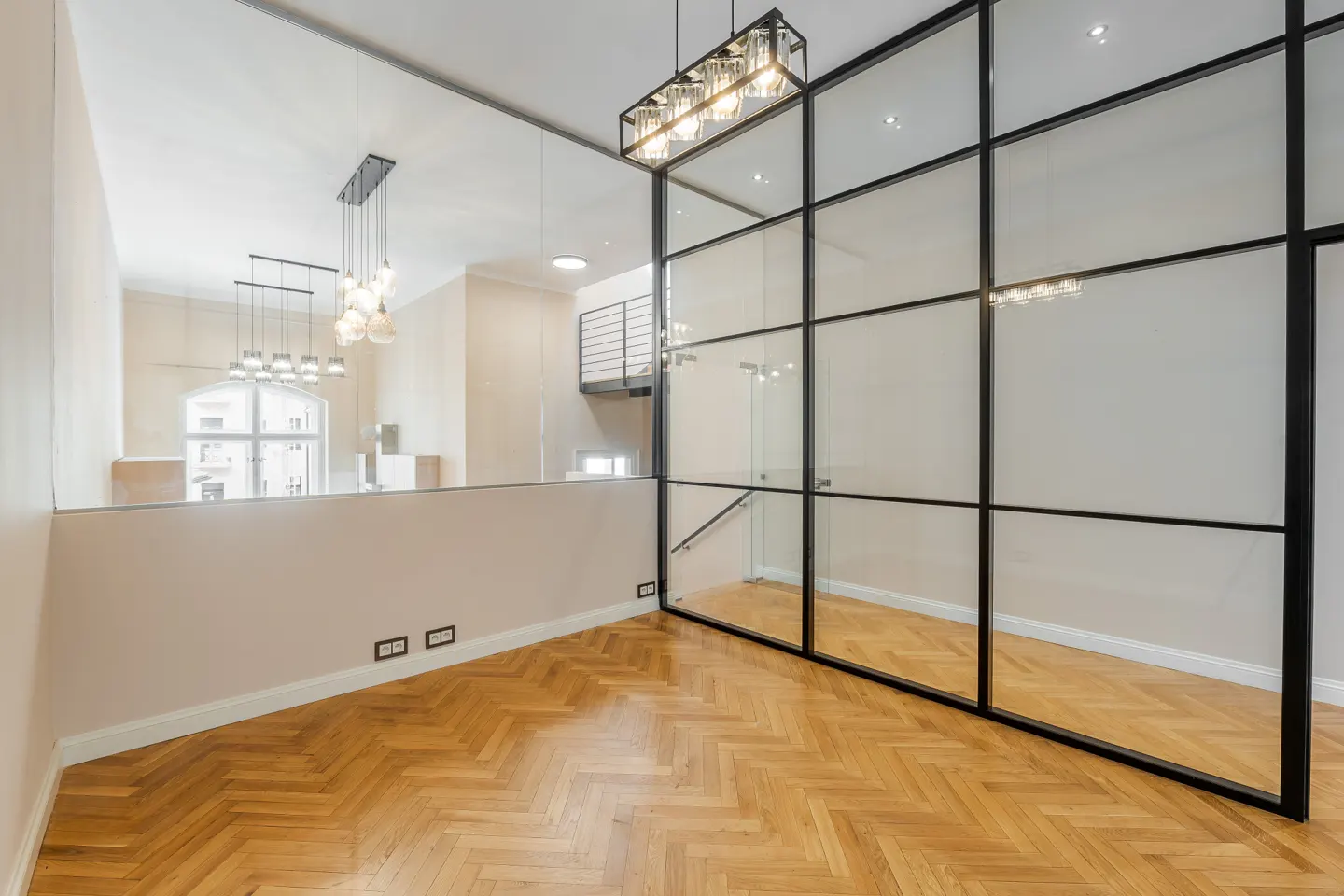 Bright room with herringbone wood floors, a black-framed glass wall, and a partial wall overlooking another room with pendant lights.