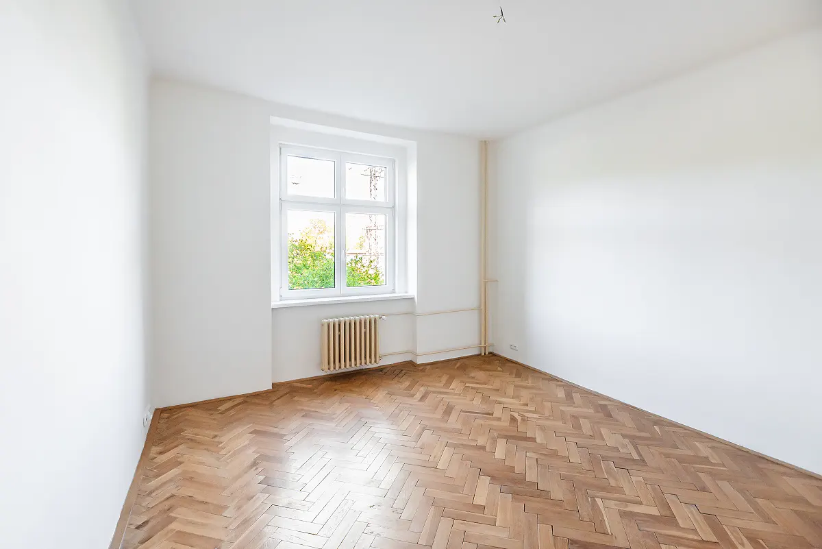 Empty room with white walls, herringbone wood flooring, a window with a view of trees, and a beige radiator.