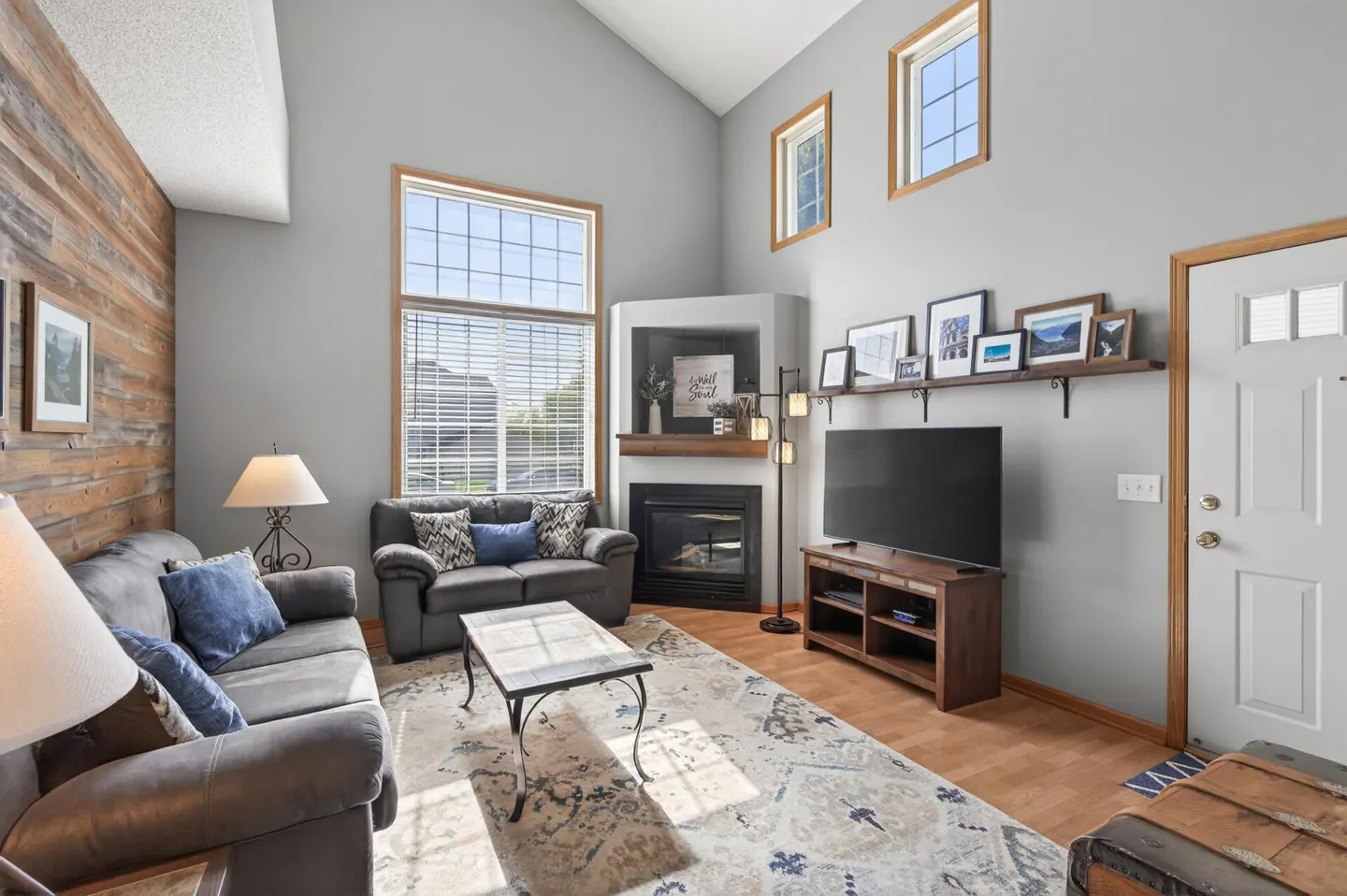 Living room with gray walls, wood accent wall, sofas, fireplace, TV, and large windows. A patterned rug sits on the wood floor.