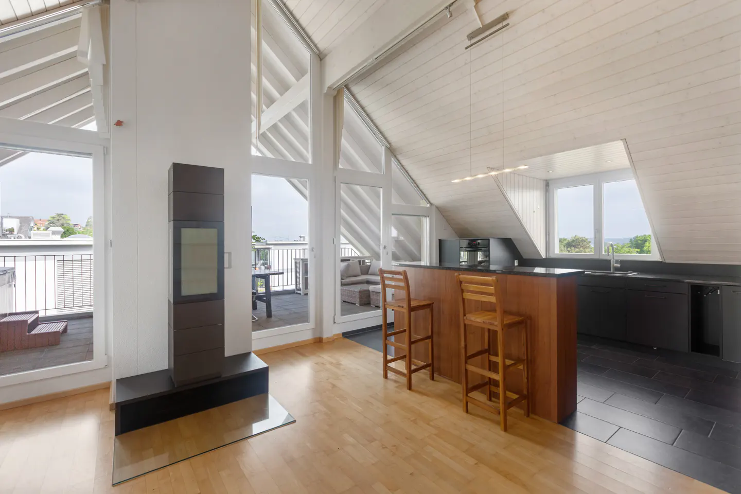 Attic apartment with wood floors, white walls, and a vaulted ceiling. A kitchen with a bar and stools is visible. Balcony with furniture is seen through large windows.