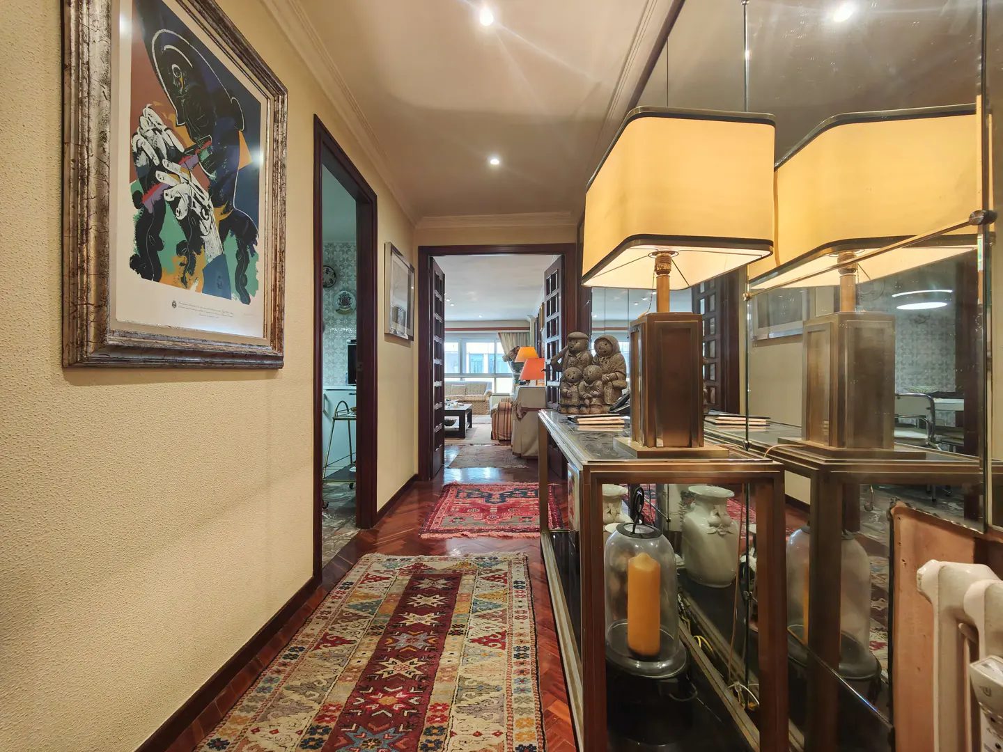 Hallway with a patterned rug, artwork, and a console table with lamps. A living room is visible in the background.