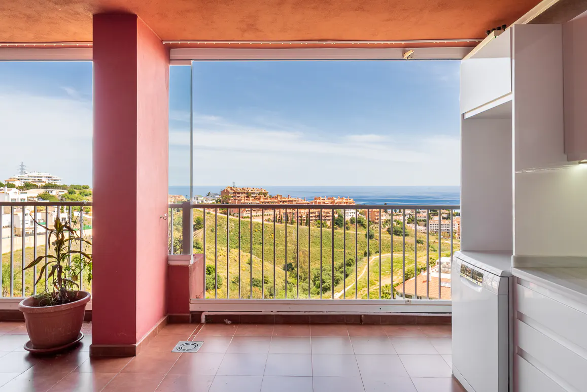 Balcony view with brown tile floor, pink column, and white cabinets. A potted plant sits near the column, and a scenic view of buildings and the ocean is visible.