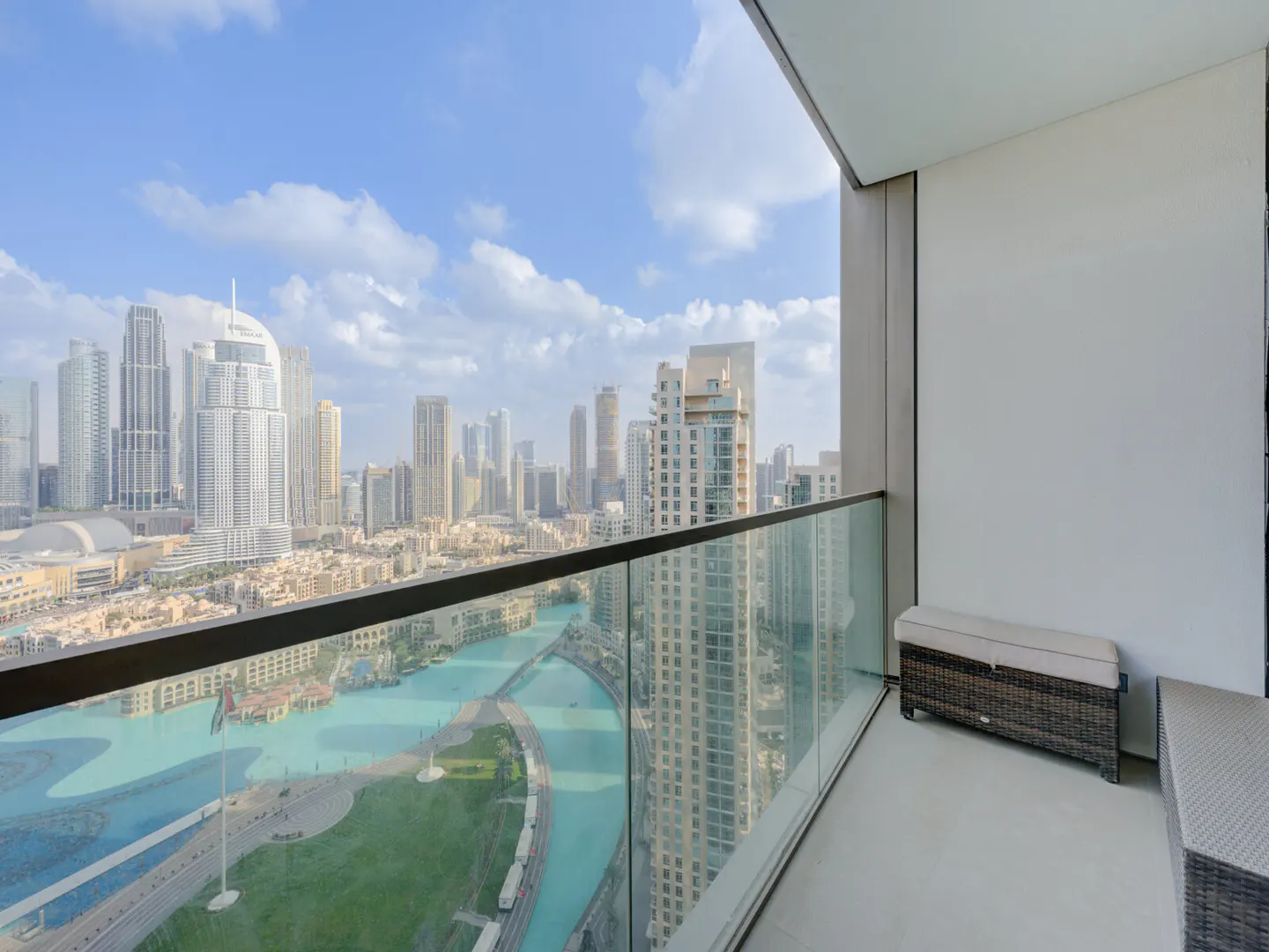 Balcony view of Dubai skyline with turquoise water feature below. Wicker bench and glass railing visible.